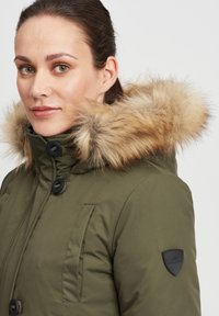 Olive green parka with faux fur hood, zip and button closure, two front pockets, and black logo patch on left sleeve. Smooth texture.
