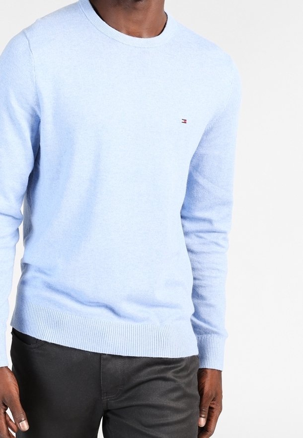 Homme portant un pull léger bleu clair à manches longues avec un petit emblème rouge, blanc et bleu sur la poitrine, et un pantalon foncé.