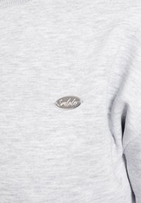 Maglione grigio chiaro realizzato in un tessuto morbido e testurizzato, con un'etichetta metallica del marchio "Smulodex" in bella vista.