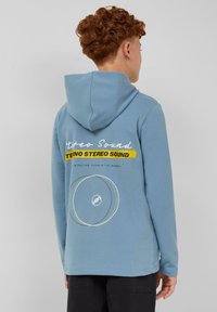 Lichtblauwe hoodie met witte tekst en gele accenten op de achterkant, met een cirkelvormig grafisch ontwerp. Zachte stofstructuur.