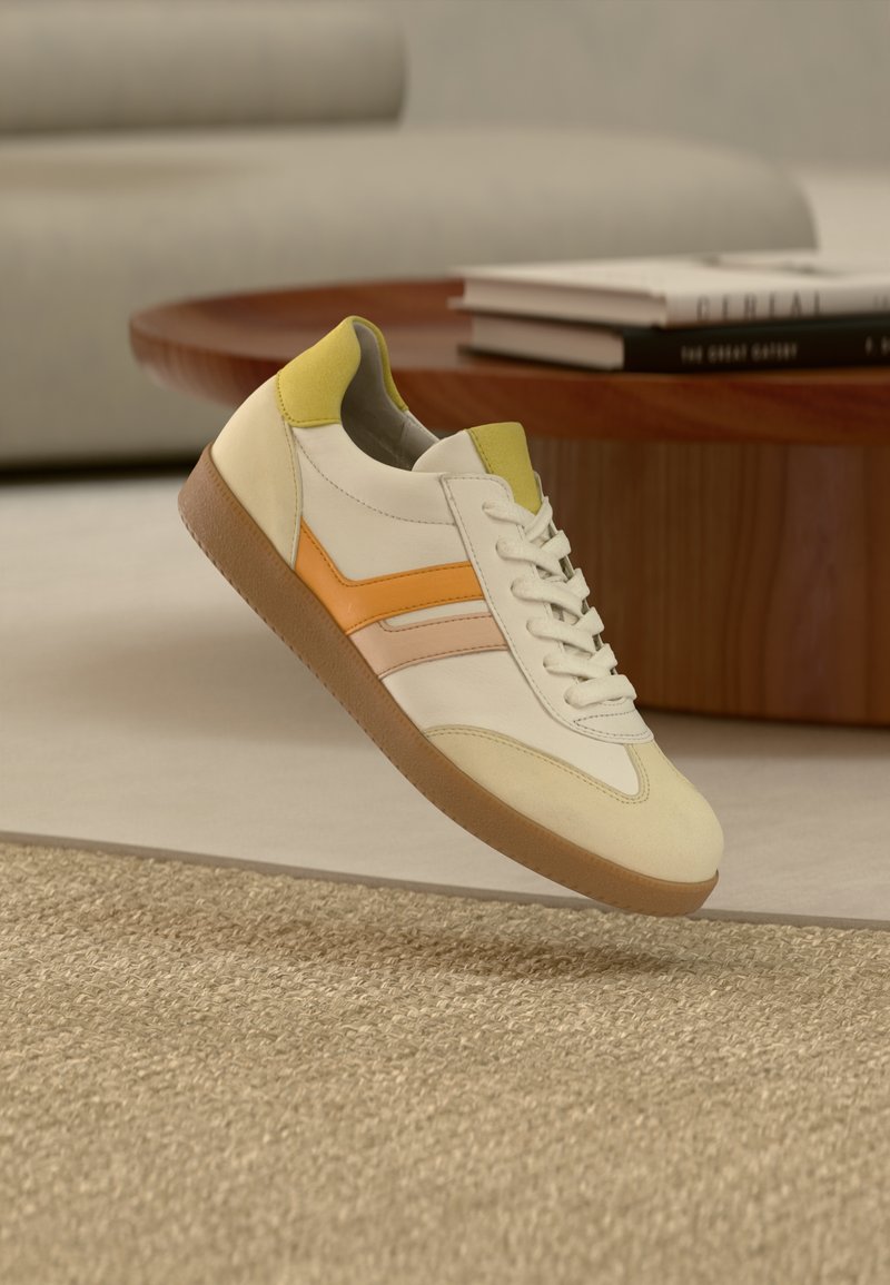 Baskets décontractées avec des accents crème, jaune et orange flottant au-dessus d'un tapis tissé beige dans un salon moderne.