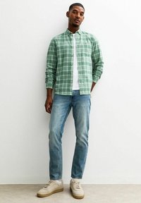 Groen geruite button-up shirt met een wit t-shirt eronder, gecombineerd met lichtblauwe jeans en beige sneakers. Malse textuur, casual pasvorm.