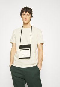 Sort og hvid crossbody taske med en lynlåslomme, lavet af glatte materialer. Tasken har et logo og justerbare stropper.
