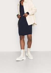 Bottes blanches à cheville avec semelles texturées, associées à une robe bleu marine et une veste-chemise en velours côtelé de couleur claire. Design simple et moderne.
