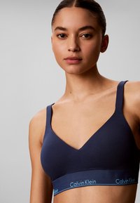 Ung kvinde med mørkt hår iført en mørkeblå Calvin Klein bralette, set forfra mod en ensartet grå baggrund.
