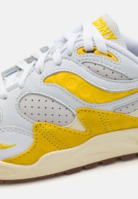 Weiße und gelbe Sneaker aus Leder und Wildleder, mit Perforationen, strukturierten Akzenten und einer Gummisohle. Die Schnürsenkel sind weiß.