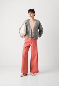 Šedý žebrovaný kardigan s kulatými knoflíky, béžový top, korálové široké kalhoty a béžové baleríny. Modelka drží krémovou kabelku.