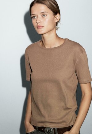 Braunes T-Shirt mit kurzen Ärmeln und rundem Halsausschnitt, glatter Textur und sichtbaren Nähten. Das Modell trägt einen dekorativen Gürtel.