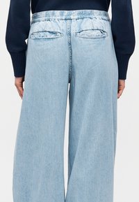 Άτομο που φοράει ανοιχτό μπλε τζιν με φαρδιά πόδια και πίσω τσέπες με καπάκια και σκούρο πουλόβερ με μακριά μανίκια, εμφανίζεται από πίσω.