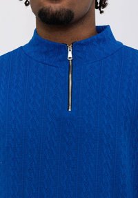 Pull-over en tricot bleu avec un col haut et une fermeture éclair demi-longueur. Présente un motif en losange sur tout le tissu pour une texture supplémentaire.