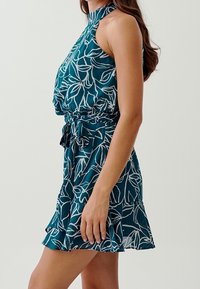 Teal halterklänning med vitt blommönster, cinchad midja och lager av volang längs fållen. Tillverkad av lätt tyg med en sl ät textur.