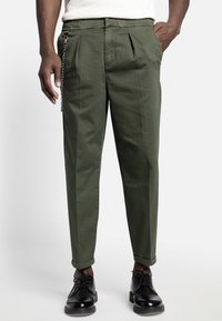 Mand i olivengrønne tapered bukser med opfoldede manchetter, sorte læder snørresko og et sølvkæde tilbehør på venstre side.