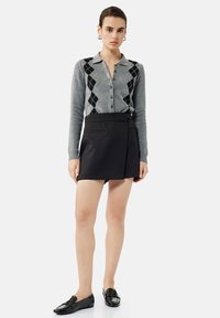 Grijze trui met zwart ruitpatroon, knoopsluiting aan de voorkant; gecombineerd met een zwarte geplooide mini rok; zwarte instapschoenen maken de outfit af.