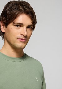 Grön bomullströja med rund hals och ribbade muddar och fåll, med en liten vit logotyp på vänster bröst. Slät textur, figursydd design.