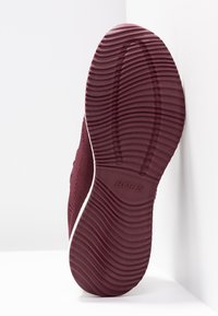 Zapato tipo mocasín de color granate con suela de goma texturizada, que presenta ranuras horizontales y detalles en blanco. El nombre de la marca "BOBS" está grabado en la suela.