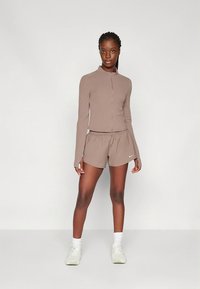 Top castanho com fecho e mangas compridas, com calções combinando. O conjunto apresenta uma textura suave e um design ajustado. Detalhe de logótipo branco nos calções.