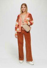 Gestreifter Strickcardigan in Pink- und Orangetönen, cremefarbenes Oberteil, braune Cordhose, weiße Sneakers und beiger Umhängetasche.