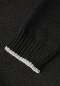 Dettaglio di un maglione nero con polsini a coste, caratterizzato da un accento bianco lungo il bordo. La texture è morbida e lavorata a maglia, mostrando un'ottima maestria artigianale.