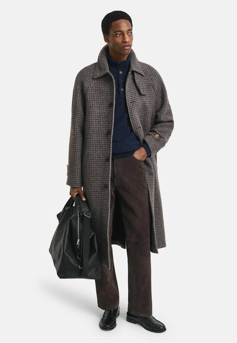 Lange houndstooth-overjas in bruin en grijs, gedragen over een marineblauwe trui. Donkerbruine broek en zwarte schoenen, met een zwarte tas.