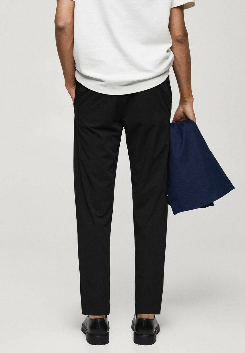 Pantalons noirs à coupe droite, portés avec un haut de couleur claire. L'individu tient une veste bleu foncé. Des chaussures noires complètent la tenue.