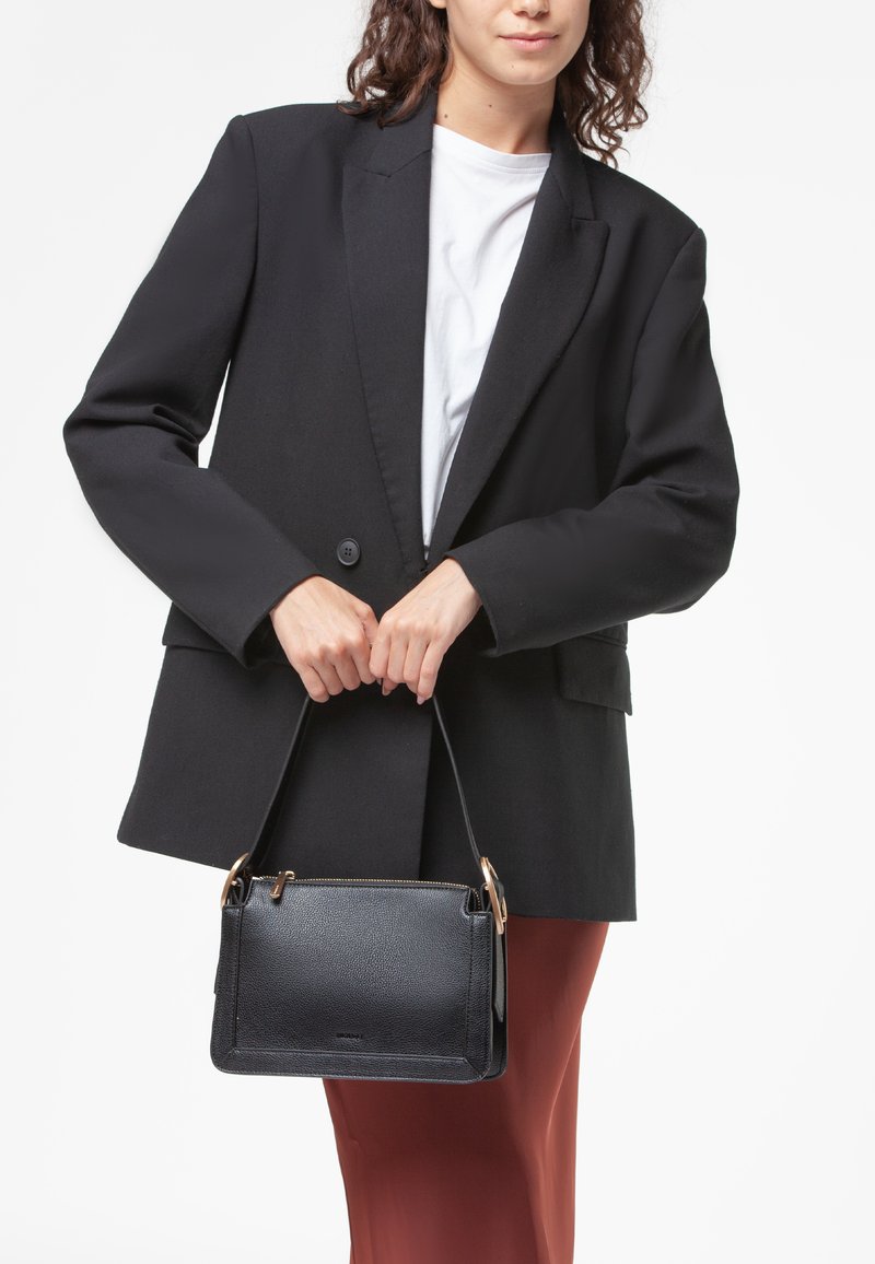 Borsa in pelle nera con forma rettangolare strutturata, dotata di dettagli in oro e chiusura con zip, esposta con un blazer nero.