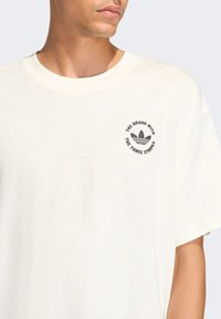 Crème kleurig t-shirt met korte mouwen, voorzien van een ronde hals en een circulair merkembleem met drie strepen in donkergrijs op de linkerborst.