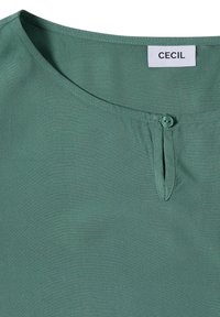 Groene blouse met een halslijn, voorzien van een kleine voorste knop en een sleutelgatdetail, met een wit label met de merknaam "CECIL".