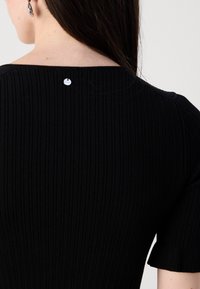 Femme portant un haut en tricot côtelé noir à manches courtes avec une petite étiquette ronde en argent centrée près de l'encolure à l'arrière.