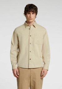 Camicia beige a maniche lunghe con chiusura a bottoni, taschino sul petto e vestibilità morbida. Realizzata in tessuto morbido con una texture liscia.