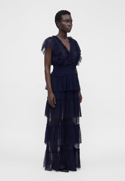 Femme debout portant une robe en dentelle bleu marine sans manches à étages avec des détails à volants et une taille cintrée sur un fond clair uni.