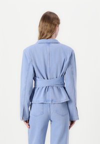 Hele sinine teksajakk, millel on struktureeritud disain, laiad kraed, tagumine õmblusdetail ja peplum ääred. Samuti on nähtavad sobivad kitsad teksad.