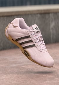 Sapatilha atlética de camurça rosa claro com atacadores brancos, três riscas castanhas e sola de borracha. Apresenta o logótipo da Adidas na língua e de lado.