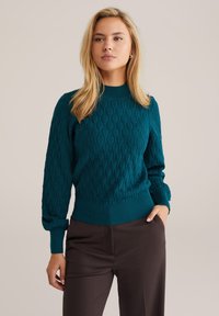Teal knitted sweater with cable pattern, ribbed crew neckline, and puffed sleeves, paired with dark brown trousers.