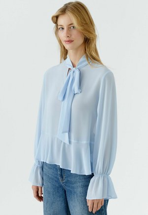 Femme blonde portant un chemisier bleu clair transparent avec un grand nœud au cou, des poignets à volants et un jean en denim bleu sur un fond blanc.