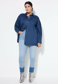 Dunkelblaues Denimhemd mit Knopfleiste, zwei Brusttaschen und langen Ärmeln. Kombiniert mit hellblauen Jeans und metallischen Knöchelstiefeln.