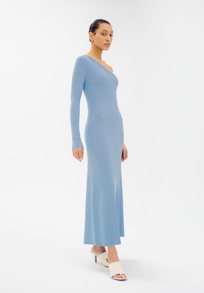 Femme portant une robe maxi en maille côtelée bleu clair à une épaule et des mules à talons ouverts blanches, se tenant devant un fond blanc.