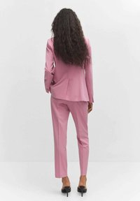 Femme aux longs cheveux bouclés debout, portant un costume rose sur mesure et des chaussures à talons noirs sur un fond clair uni.