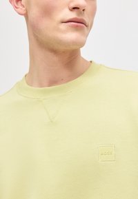 Άνδρας που φοράει ανοιχτό κίτρινο φούτερ με στρογγυλή λαιμόκοψη και μικρό ανάγλυφο λογότυπο "BOSS" στο στήθος, στραμμένος ελαφρώς προς τα δεξιά, με ουδέτερο φόντο.