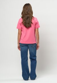 Ženska z dolgimi rjavimi lasmi, obrnjena proti steni, oblečena v roza majico s kratkimi rokavi in modre kavbojke na enostavni podlagi.