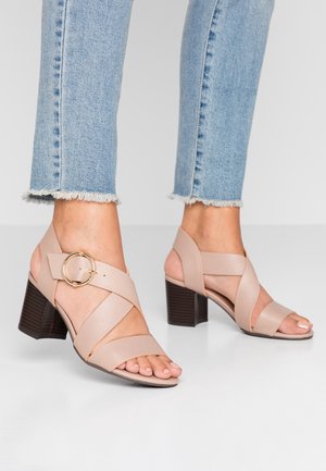 Beiget korkokengät, joissa on ristikkäiset remmit, avoin varvas ja kullanvärinen pyöreä koriste. Niissä on paksu puukuvioitu lohkokorko.