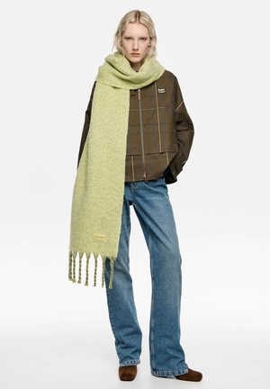 Mujer con chaqueta de cuadros verdes sobredimensionada, gran bufanda verde claro, vaqueros azules y zapatos marrones, posando frente a un fondo blanco.