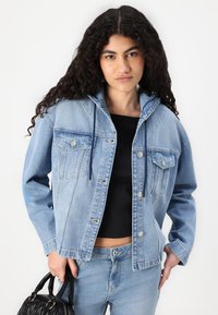Svetlo modra denim jakna s kapuco, sprednjimi žepi in srebrnimi gumbi. V kombinaciji s črnim zgornjim delom in svetlo modrimi kavbojkami, drži črno torbico.