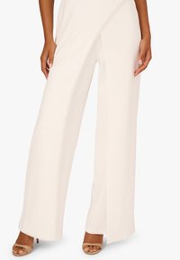 Kalhoty s širokými nohavicemi z měkké bílé látky, zdobené po straně drobnými kulatými ozdobami, doplněné o tělové podpatky.