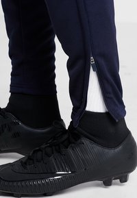 Černé kopačky nošené s černými ponožkami a tmavě modrými sportovními kalhotami s částečně rozepnutým zipem na kotníku, který odhaluje bílé ponožky.