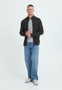 Karbonisert teksturert zip-up jakke over en grå t-skjorte, kombinert med lyseblå jeans og grå sneakers. Modellen står med hendene foldet.