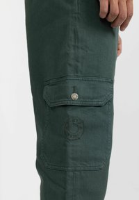 Green cotton cargo pants feature a side pocket with a snap closure and embossed logo detail. The fabric has a smooth texture and sturdy design.
