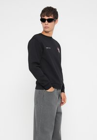 Fekete pamut pulóver hímzett logóval a mellkason, szürke, bőszárú farmernadrággal párosítva. A modellen fekete napszemüveg van, kezei a zsebében.