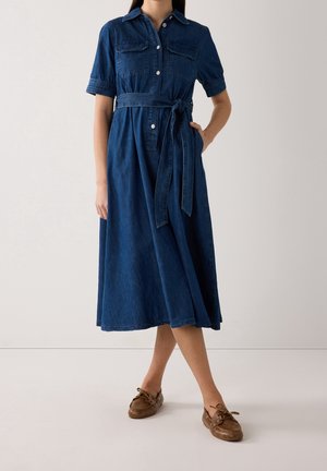 Kvinde iført en blå denim midi skjortekjole med korte ærmer, knapper foran, brystlommer, et bindebælte i taljen og brune mokkasinsko.