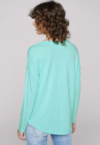 Mintgrön långärmad topp med rundad halsringning, mjuk textur och krökt nederkant. Har en centralseamsdetalj längs ryggen.