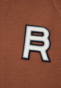 Tessuto a maglia marrone con una grande lettera "R" ricamata in bianco e bordatura blu navy. Superficie texturizzata e cuciture visibili.